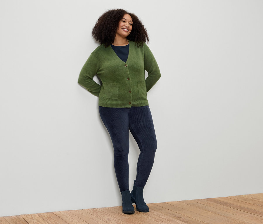 Kıvırcık saçlı bir kadın duvara yaslanmış. Yeşil bir hırka, mavi tayt ve bot giyiyor.