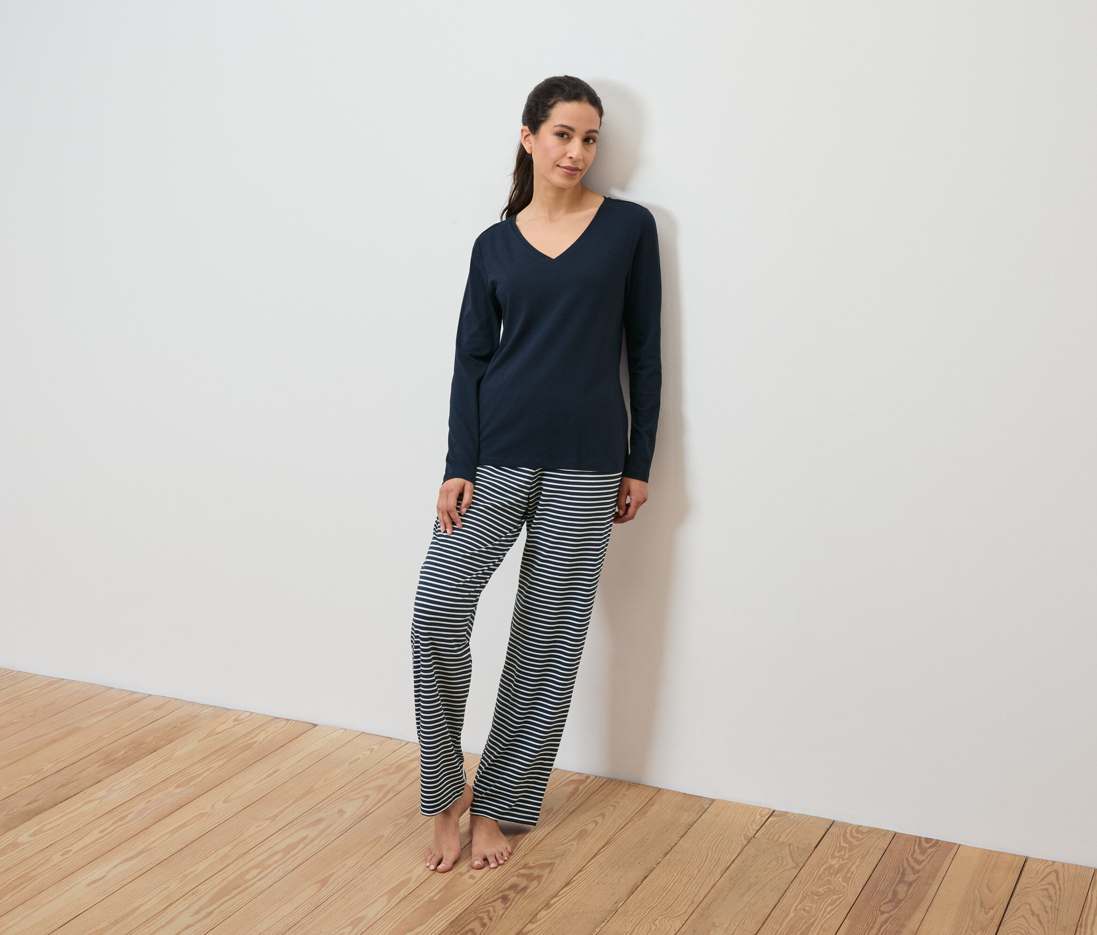 Lacivert pijamalık ve çizgili pantolon giyen bir kadın beyaz bir duvara yaslanıyor.