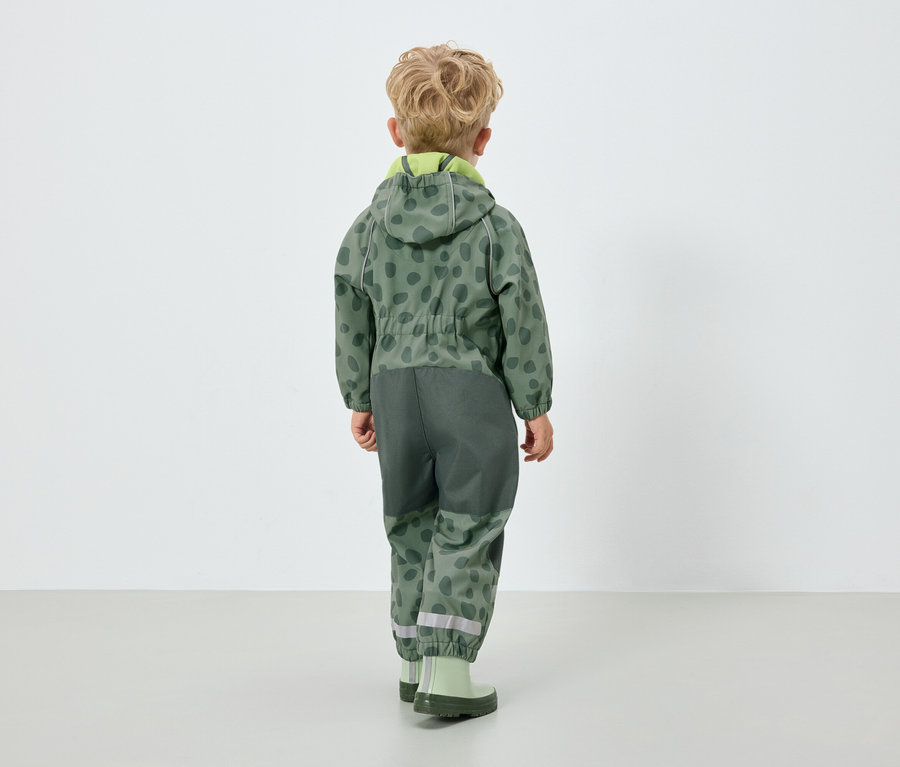 Kapüşonlu yeşil tulum giyen bir erkek çocuk, kameraya arkası dönük.