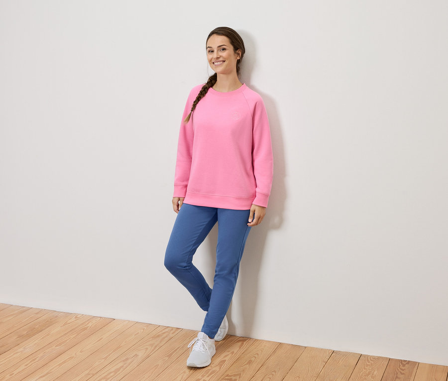 Örgülü saçlı, pembe sweatshirt, mavi pantolon ve beyaz ayakkabılar giyen bir kadın duvara yaslanmış.