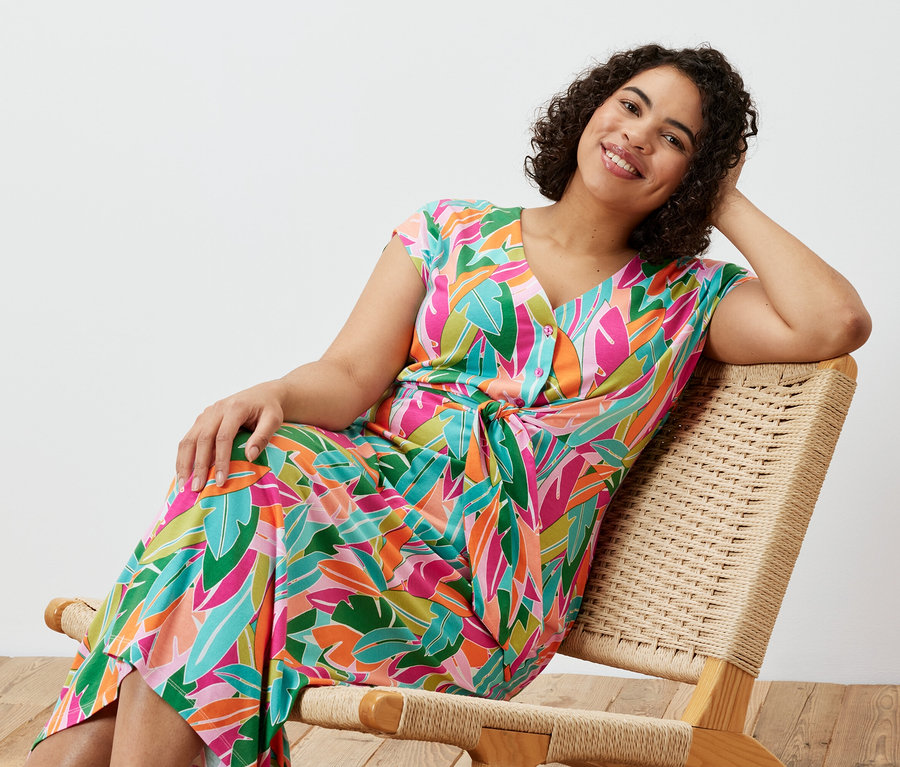 Bir kadın sandalyede oturuyor ve düğümlü jarse bir elbise giyiyor.