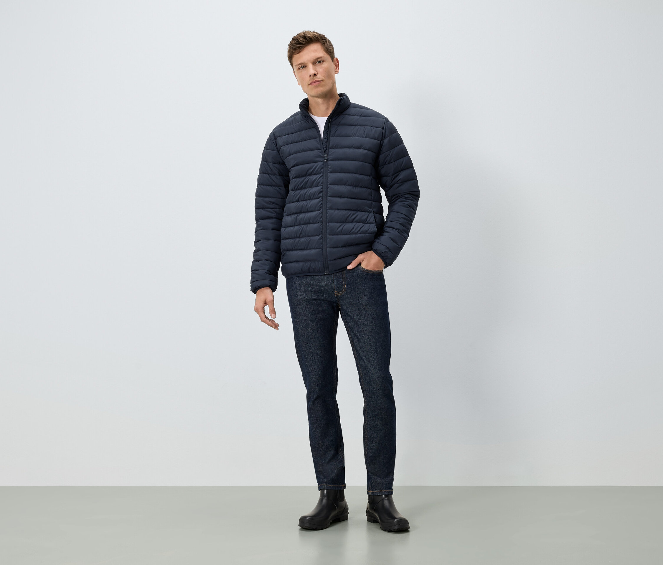 Bir adam eli cebinde duruyor. Üzerinde mavi bir ceket, kot pantolon ve siyah botlar var.