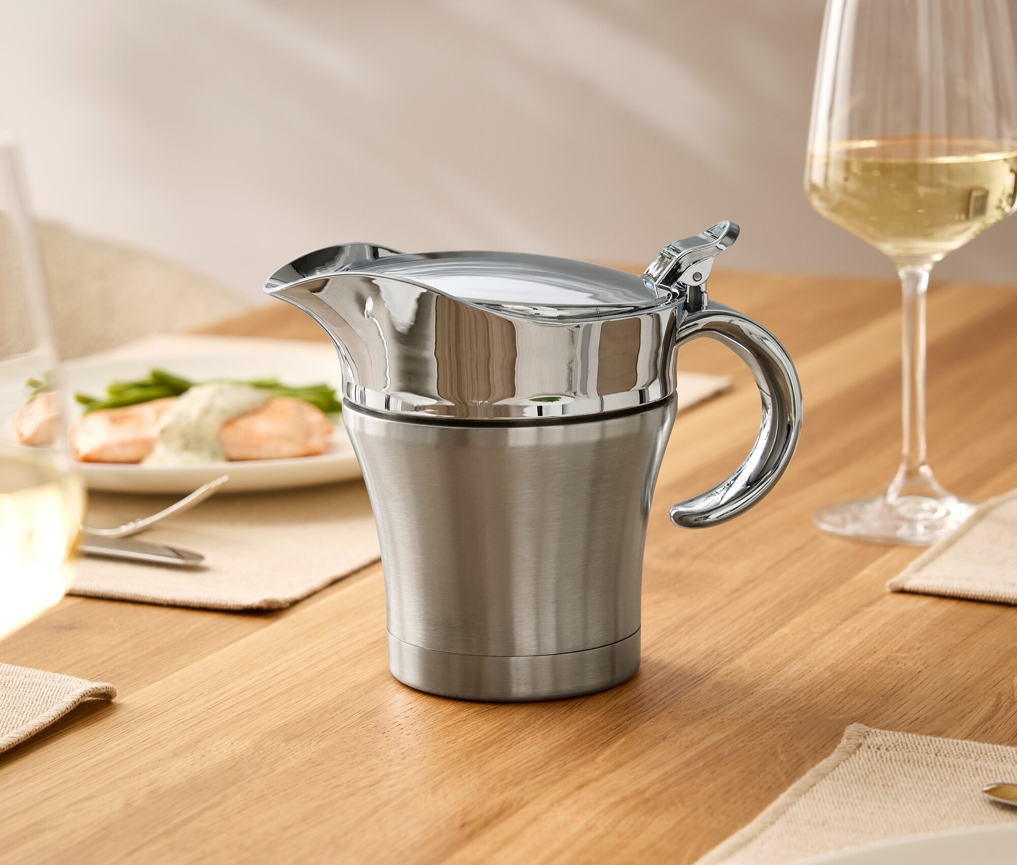 Ahşap bir masanın üzerinde kapaklı paslanmaz çelik sosluk, bir bardak şarap ve yemekli tabaklar duruyor.