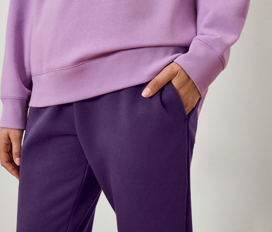 Lila bir sweatshirt ve koyu mor eşofman altı giyen bir kişinin detayı, bir eli cebinde.