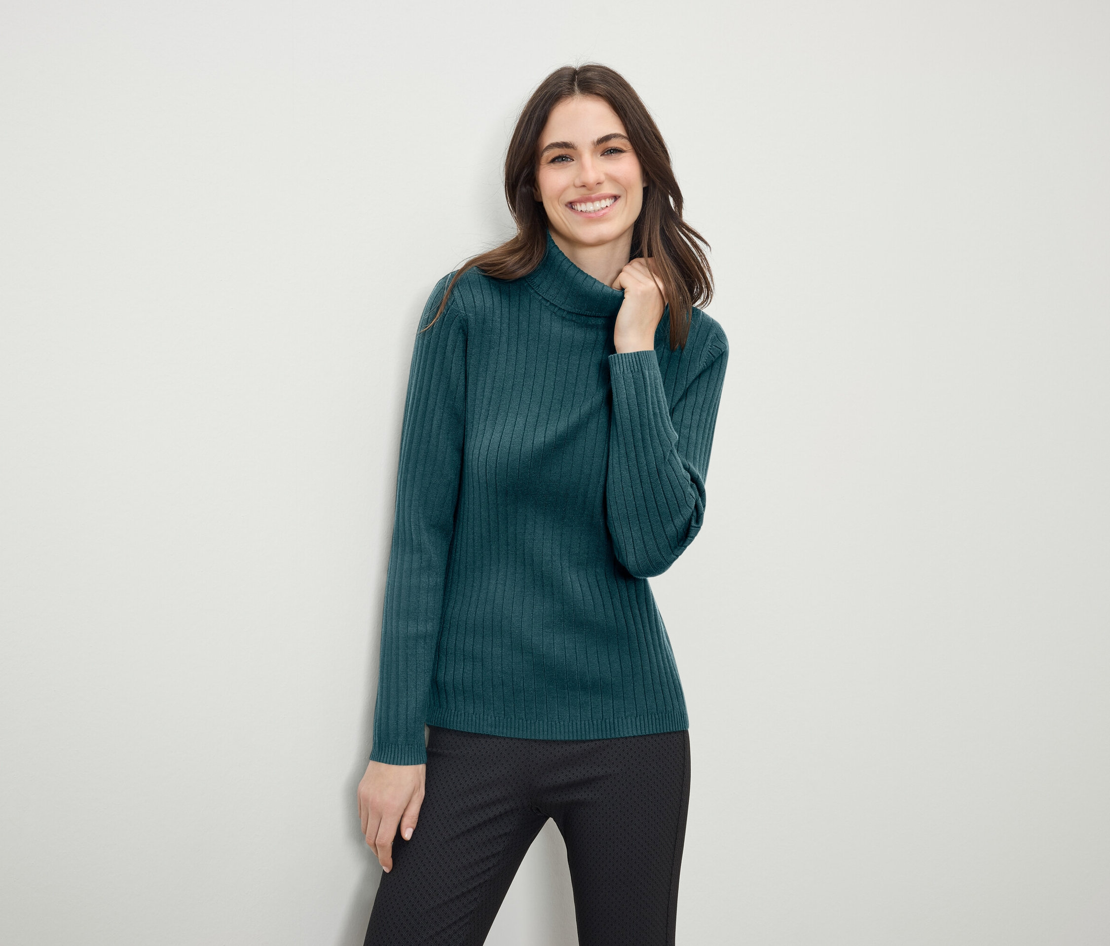 Gülümseyen bir kadın, yeşil boğazlı kazak ve siyah pantolonla poz veriyor.