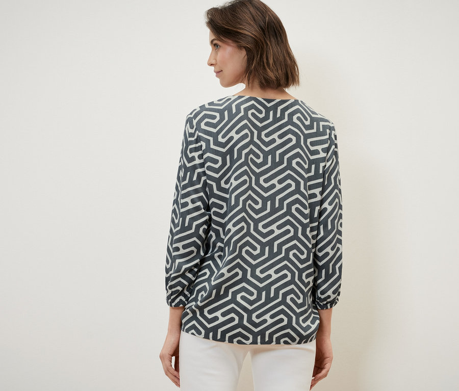 Kadın kameraya arkası dönük, kırışık görünümlü bir bluz ve beyaz pantolon giyiyor.