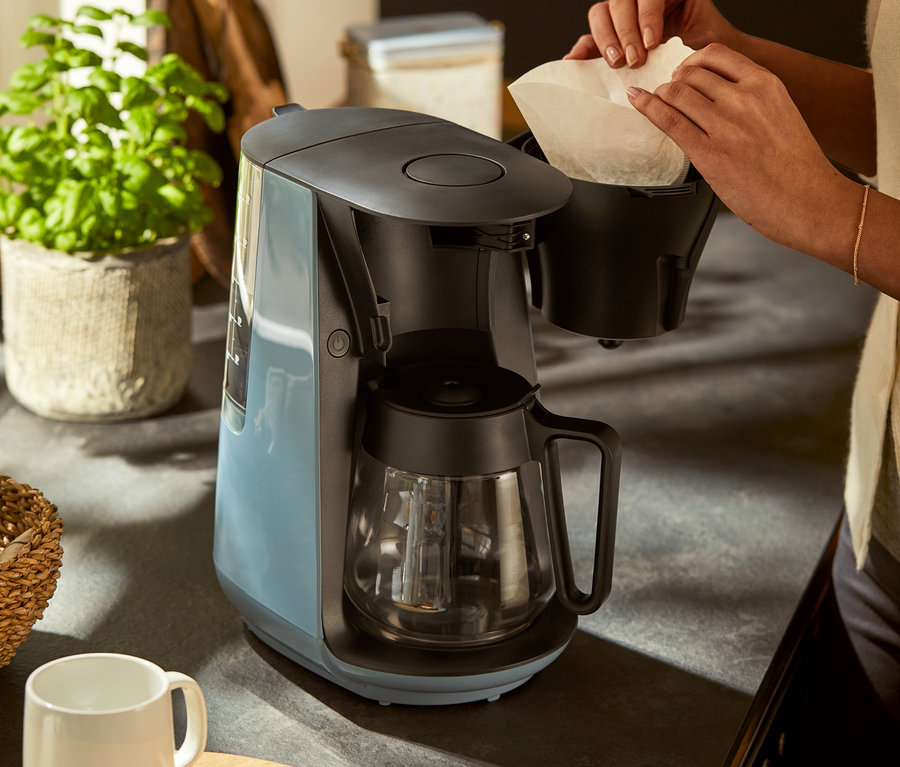 Kadın, gri mavi renkli »Let's Brew« filtre kahve makinesine kağıt filtre yerleştiriyor.