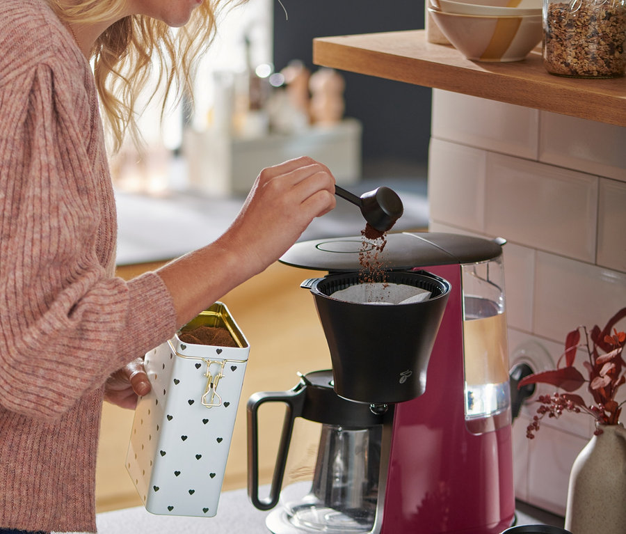 Sarışın saçlı bir kadın, Berry rengindeki »Let's Brew« Filtre Kahve Makinesine kahve döküyor.