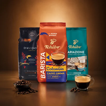 Üç kahve paketi dik duruyor: Davidoff Origins Brazil Espresso, Tchibo Barista Colombia Caffè Crema ve Tchibo Ariazione Coffee-Milk-Specialties. Ön planda bir kase kahve çekirdeği ve bir fincan kahve var.