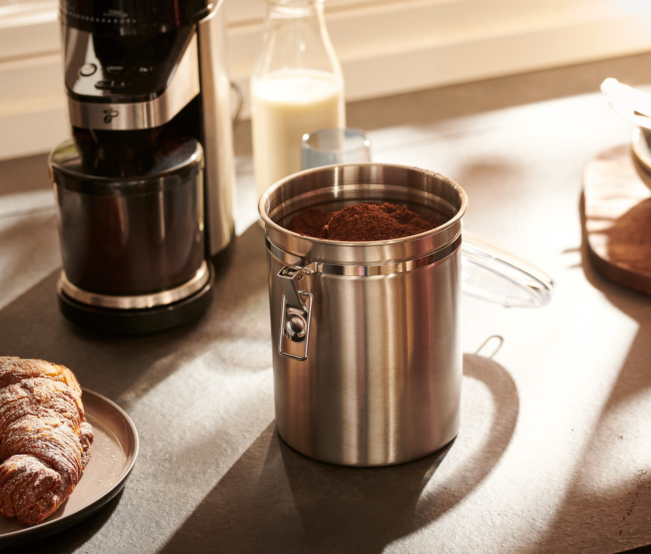 Paslanmaz çelikten bir aroma kutusu, bir kahve makinesi ve kruvasanlı bir tabağın yanında bir mutfak tezgahında duruyor.