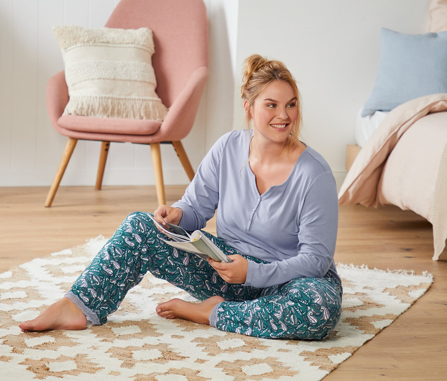 Kadın yerde halının üzerinde oturmuş kitap okuyor. Üzerinde tamamı baskılı pijama takımı var.