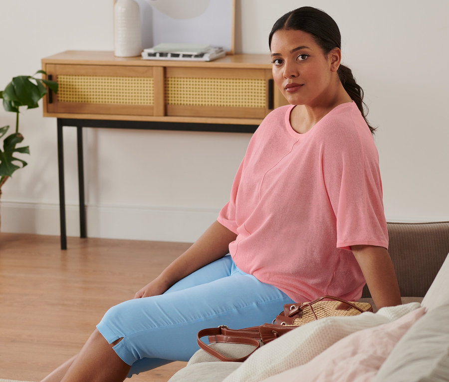 Bir kadın koltukta oturuyor ve üzerinde ince triko karışımlı bir kazak, açık mavi kısa paçalı bengalin pantolon ve espadril var. Koltuğun üzerinde büzgülü çanta duruyor.