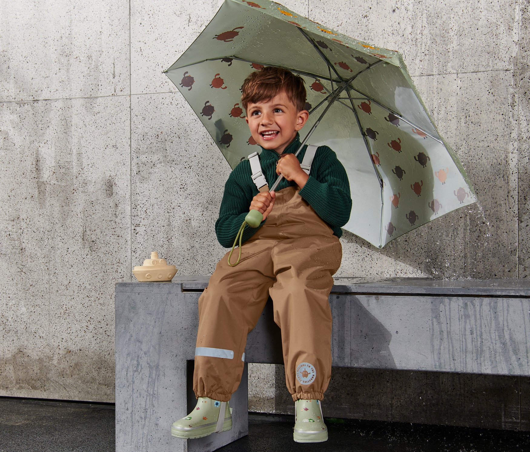 Şemsiyesiyle oturan bir çocuk, üzerinde kahverengi termal yağmur pantolonu var.