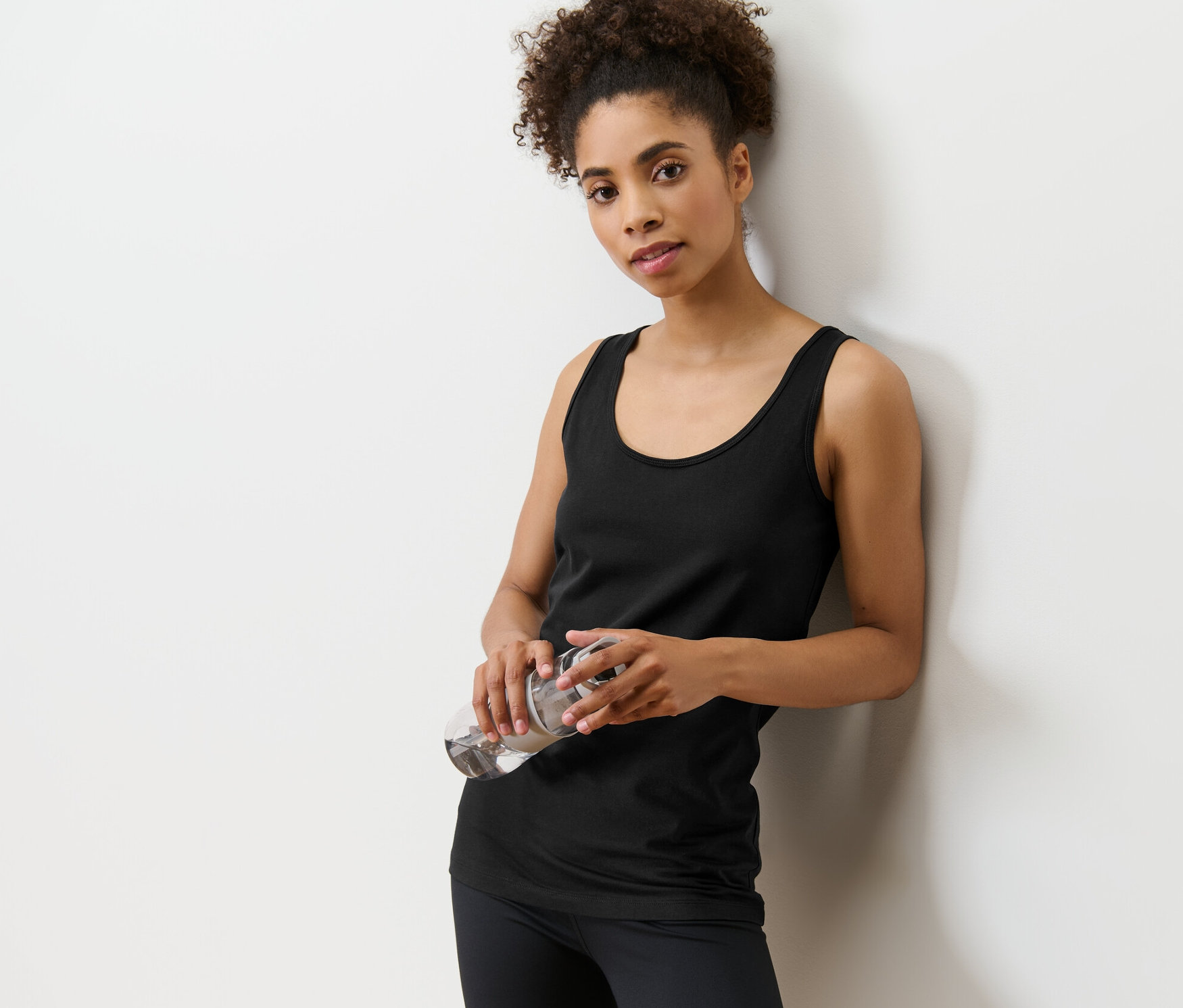 Duvara yaslanmış, elinde su şişesi tutan kadın. İki parçalı atlet setinden siyah bir atlet giyiyor.
