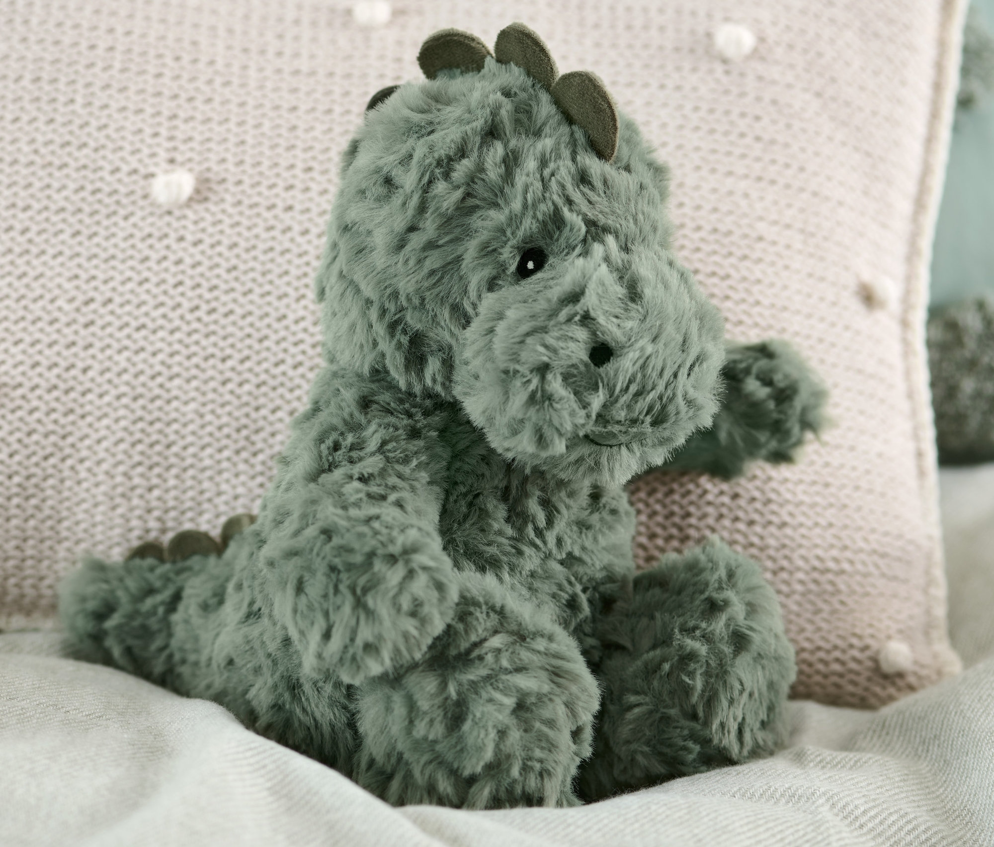 Pelüş dinozor bej bir yastığın yanında yatakta oturuyor.