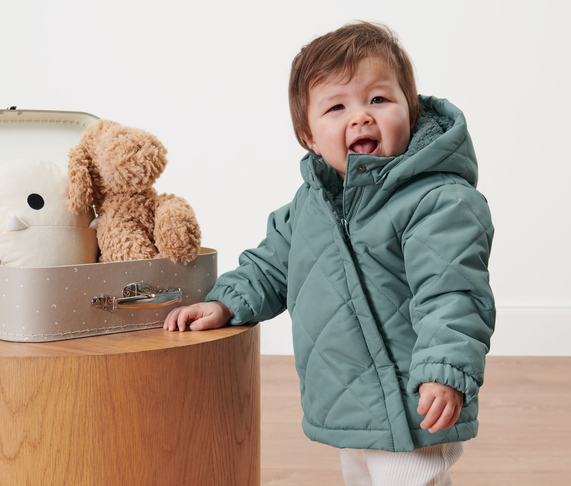 Küçük çocuk kapitone montuyla oyuncakların olduğu bavulun yanında duruyor.