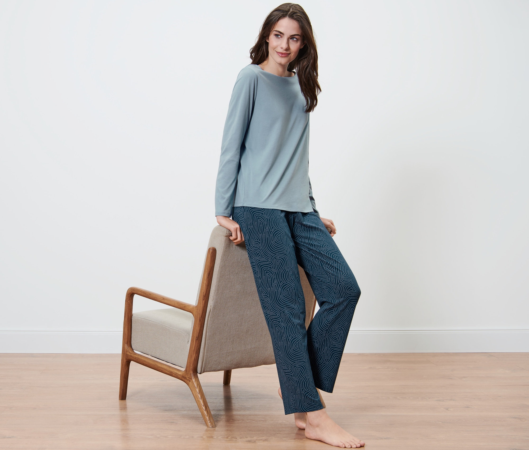 Bir kadın sandalyeye yaslanmış oturuyor ve mavi renkli jarse pijama takımı giyiyor.