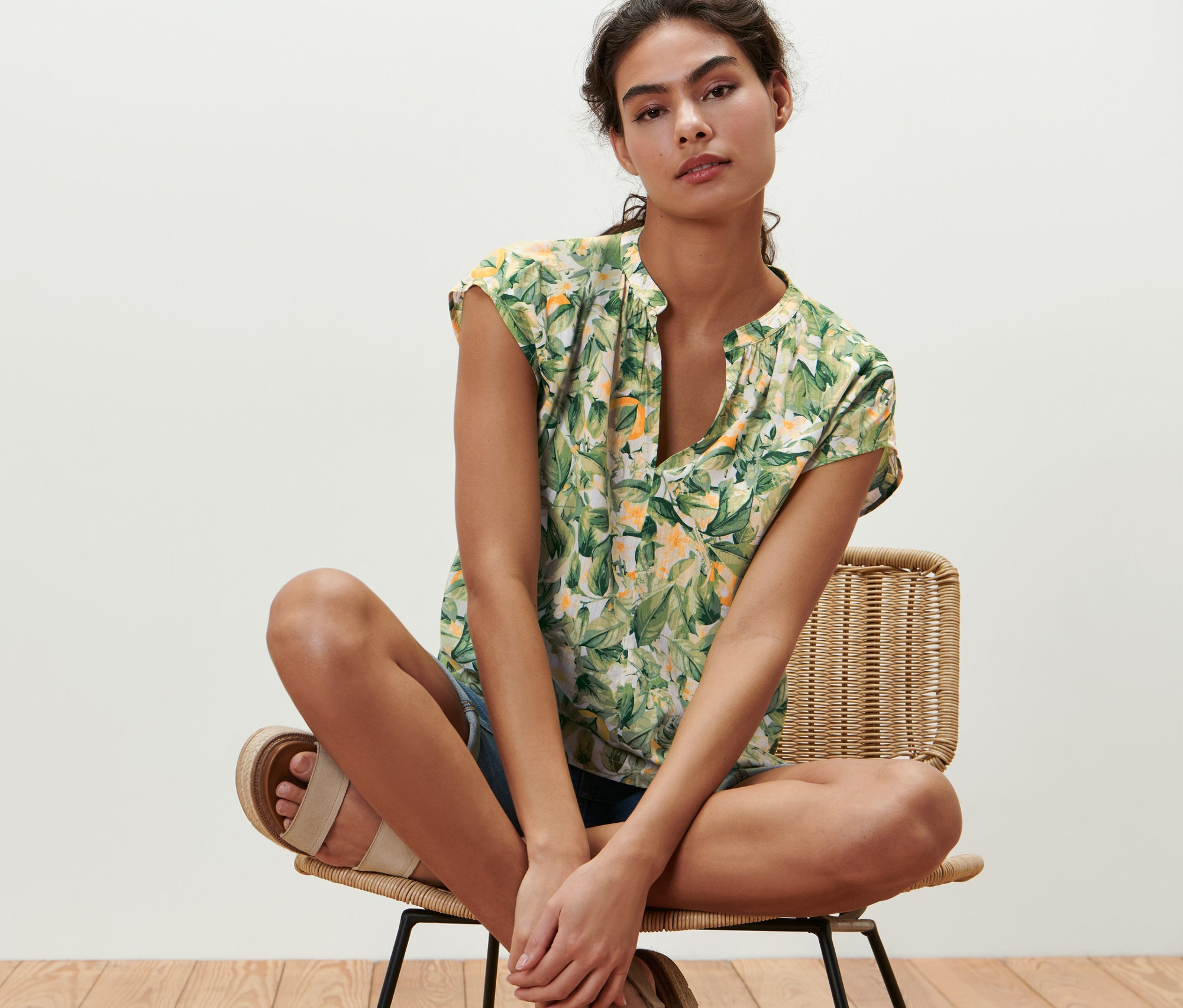 Kadın sandalyede oturuyor ve baskılı kısa kollu bir bluz giyiyor.