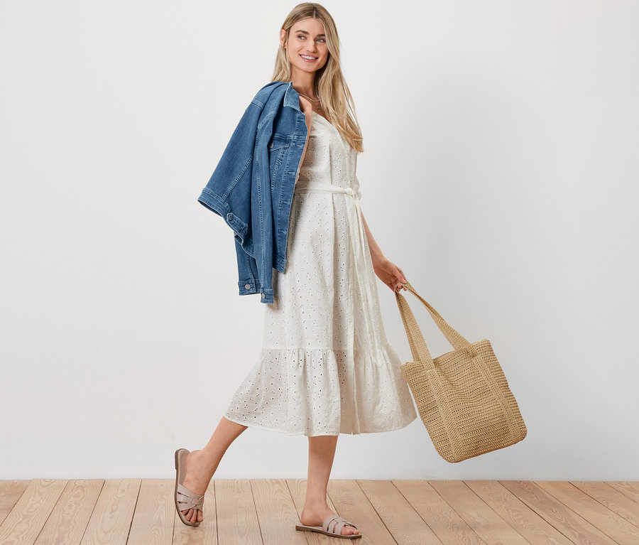 Omuzunda Kot Ceket, elinde Hasır Görünümlü Alışveriş Çantası ve üzerinde Brodeli Bluz Elbise olan bir kadın yürüyor.