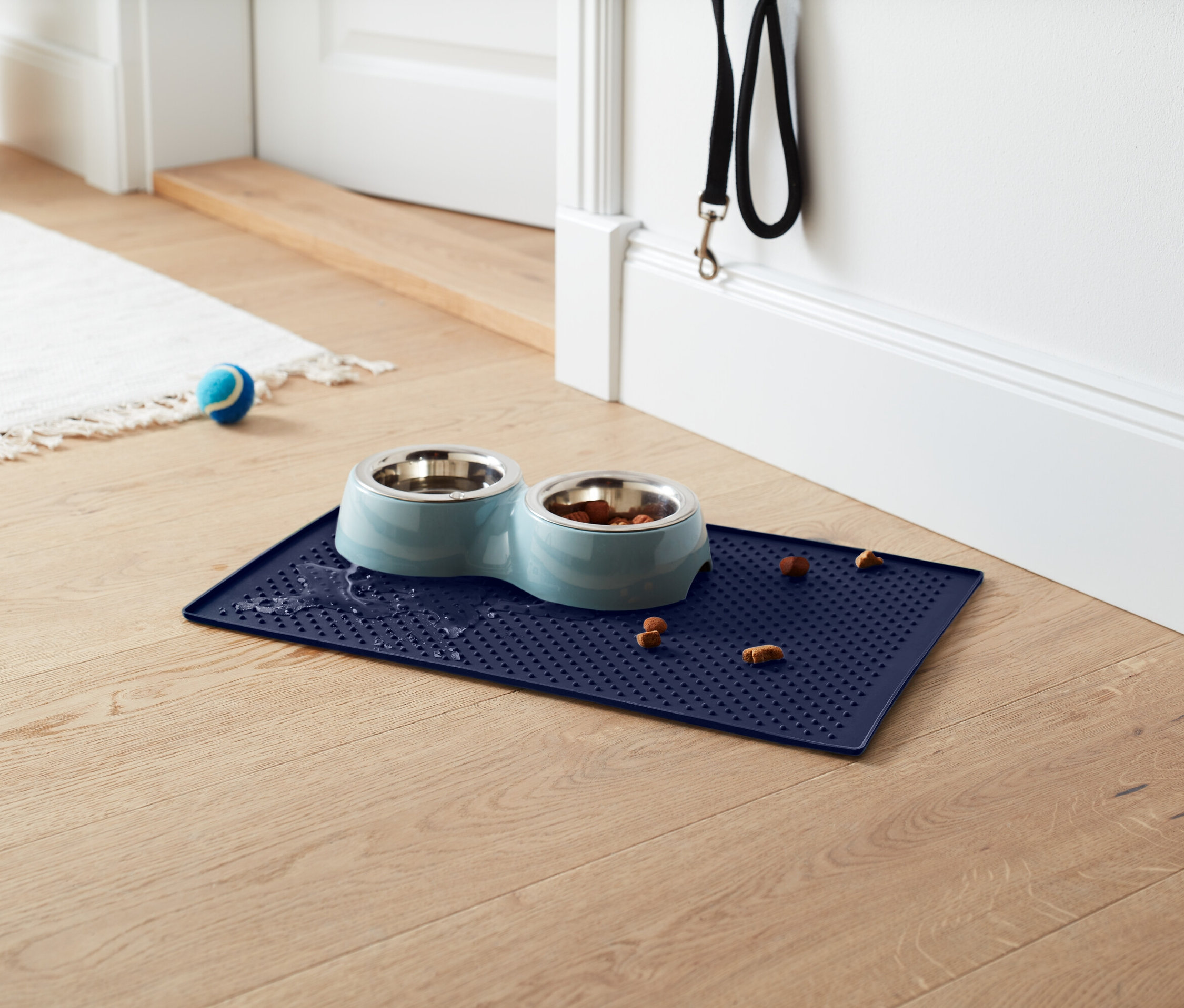 Mavi mama kabı altlığı üzerinde mama kapları var, bir tanesi mama dolu, altlıkta ve etrafında mama parçaları var. Arka planda beyaz bir halı, mavi bir top ve siyah bir tasma bulunuyor.