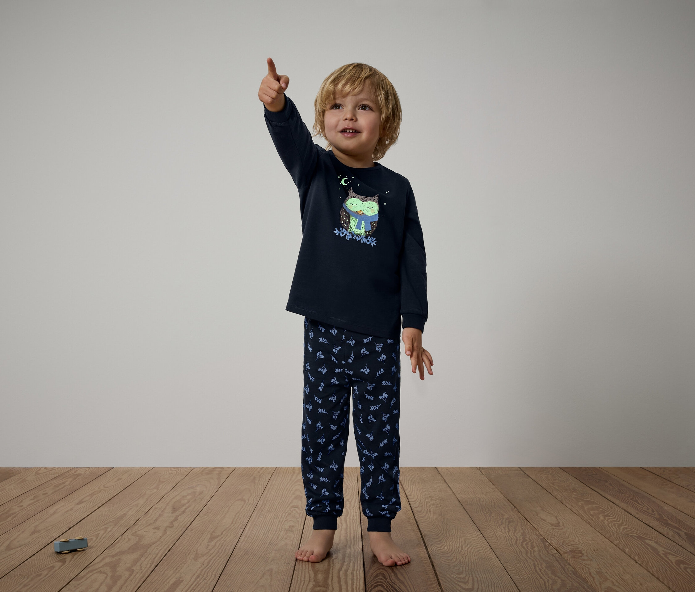 Baykuş desenli lacivert pijamalı bir çocuk parmağıyla yukarıyı işaret ediyor.