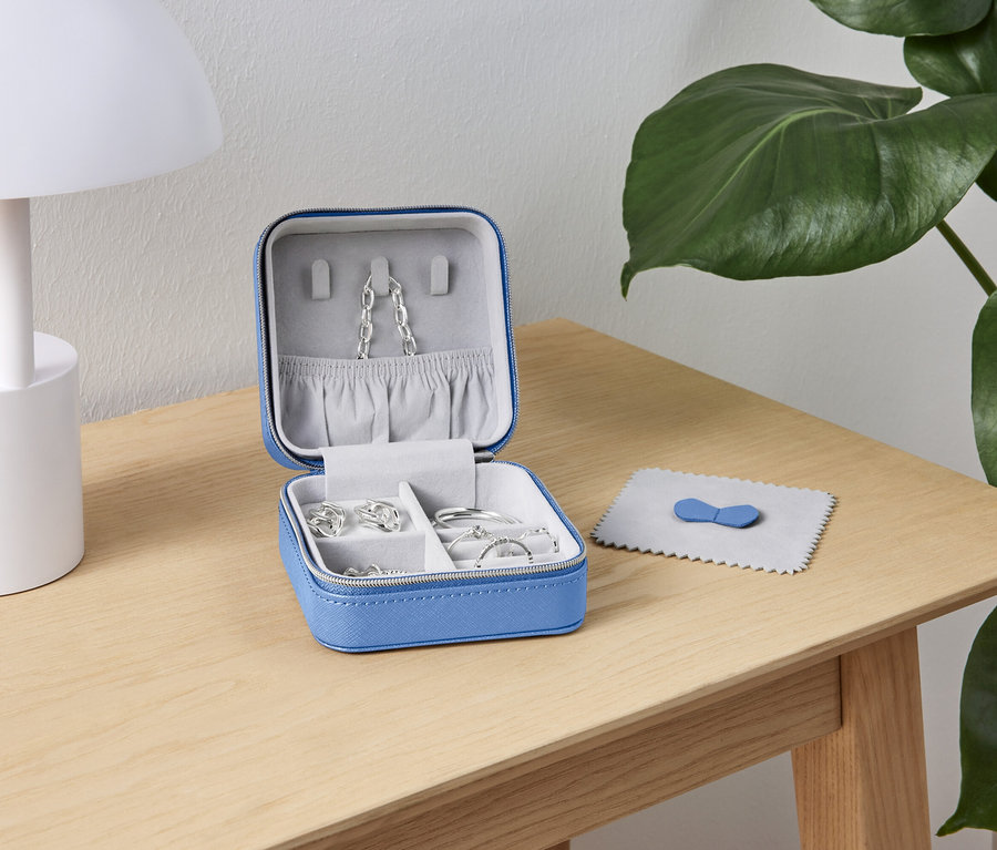 Açık mavi mücevher kutusu, gümüş takılarla birlikte ahşap bir masanın üzerinde beyaz bir lamba ve yeşil bir bitkinin yanında duruyor.