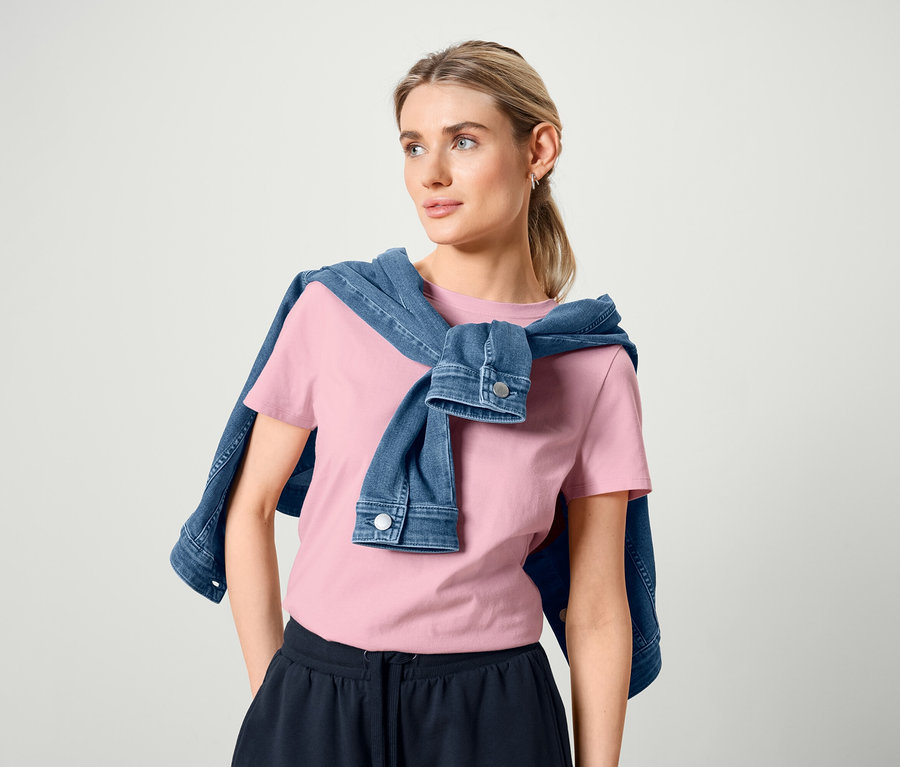 Omuzlarında kot ceket olan kadın, pembe basic tişört ve bermuda eşofman altı giyiyor.