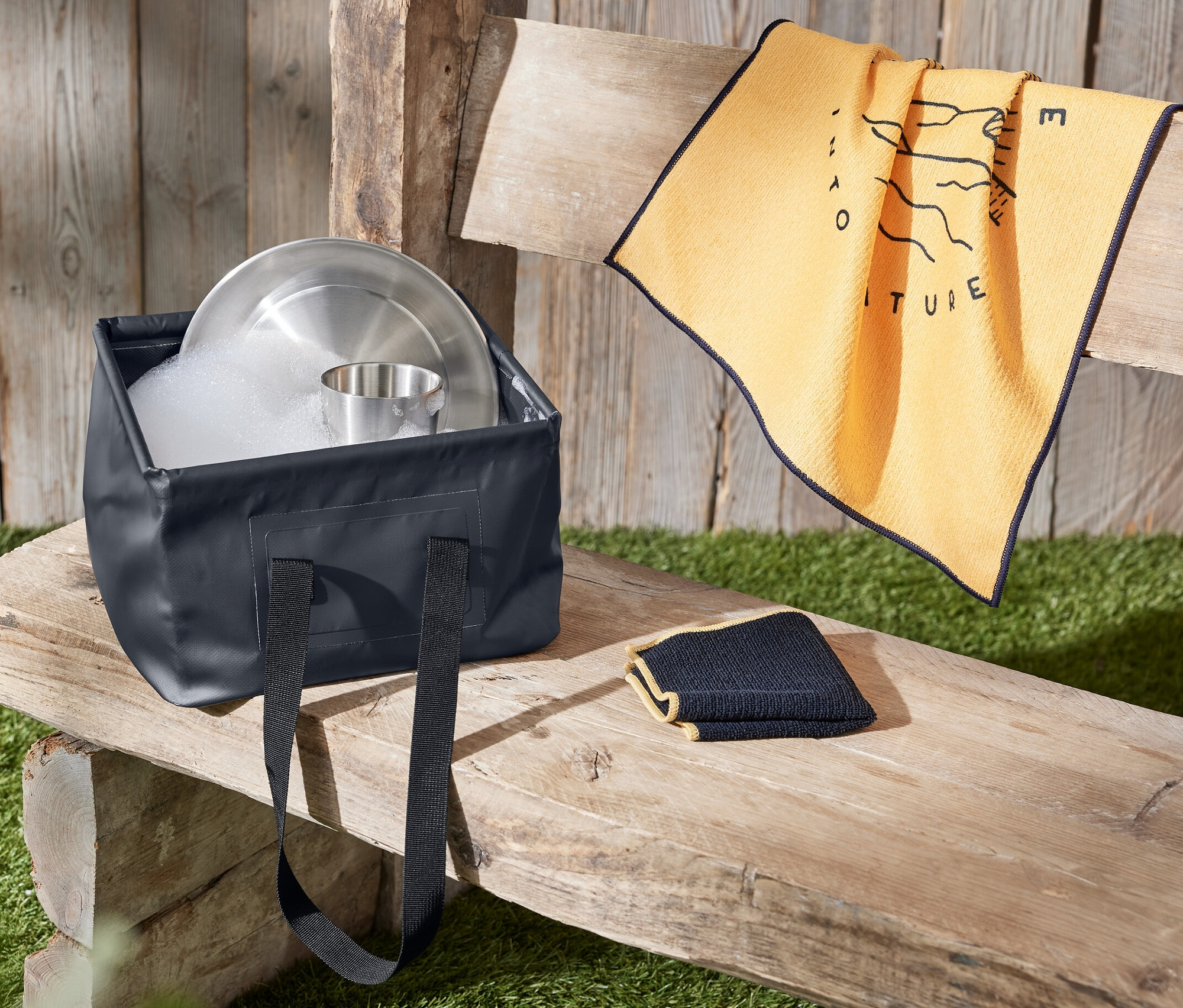 Katlanabilen Bulaşık Kabı su ve bulaşıklarla dolu halde tahta bir bank üzerinde bir havlunun yanında duruyor.