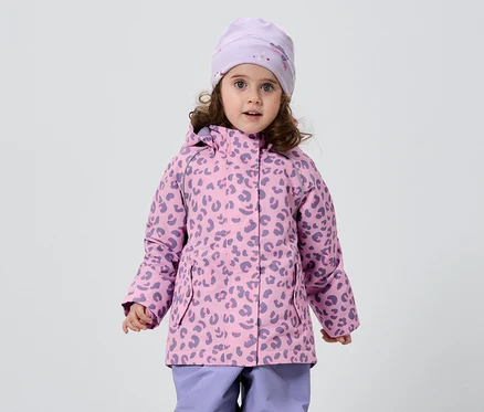 Kıvırcık saçlı küçük bir kız, beyaz bir arka plan önünde leopar desenli pembe-mor bir ceket, uyumlu bir şapka ve mor pantolon giyiyor.