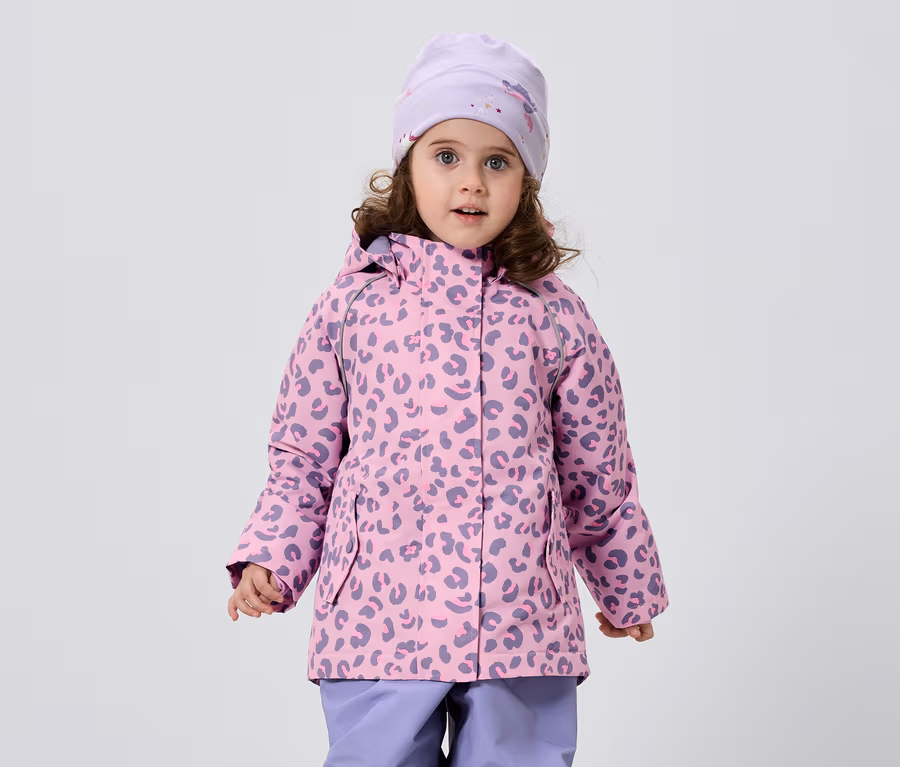 Kıvırcık saçlı küçük bir kız, beyaz bir arka plan önünde leopar desenli pembe-mor bir ceket, uyumlu bir şapka ve mor pantolon giyiyor.