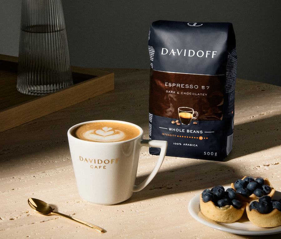 Bir fincan Davidoff Café latte sanatı, bir paket Espresso 57, yaban mersinli tartlar, altın bir kaşık ve bir sürahi masa üzerinde.