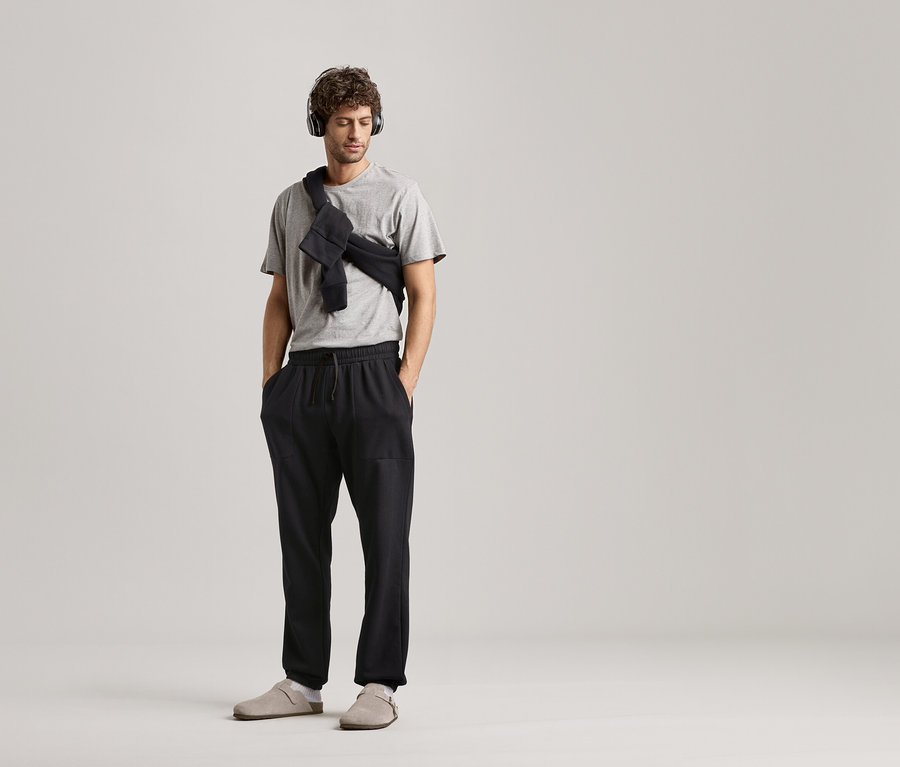 Nötr bir arka planda kulaklıklı, gri tişörtlü ve siyah eşofmanlı adam.