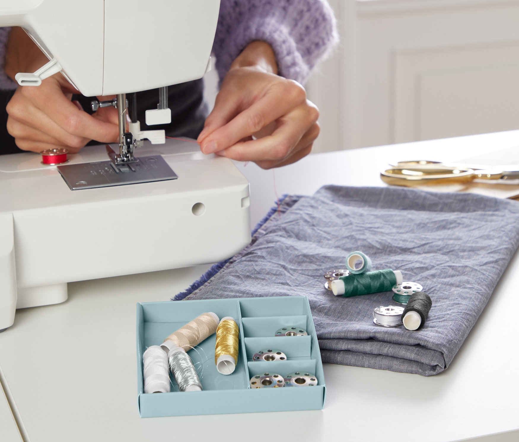 Kadın dikiş makinesine iplik geçiriyor. Dikiş İpliği Seti masanın üzerinde mavi kutuda duruyor.