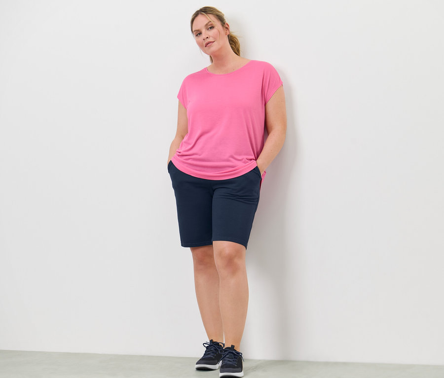 Kadın elleri şort cebinde duruyor. Üzerinde pembe bir spor tişörtü var.