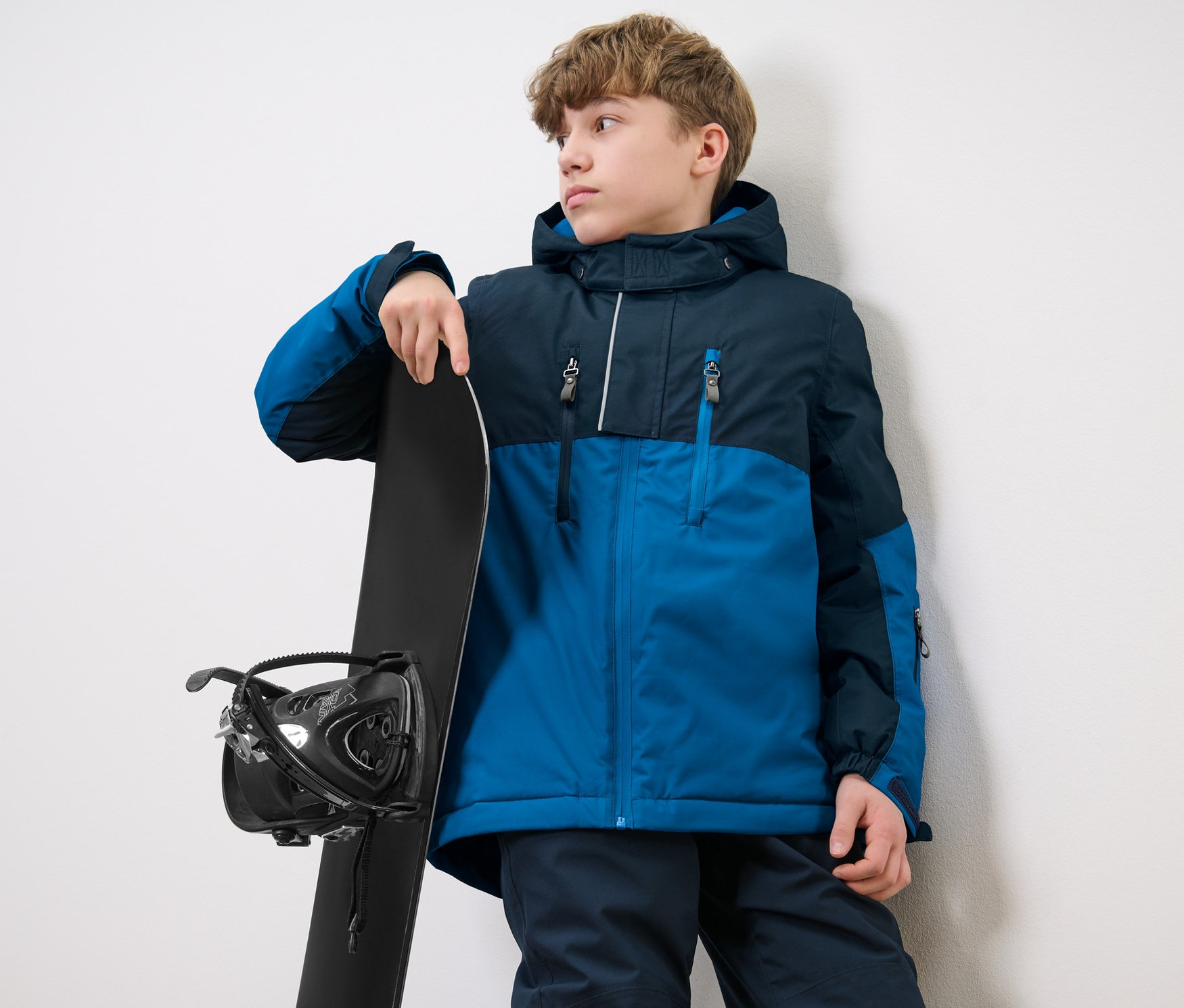 Çocuk kayak montu giyen bir çocuk snowboardun yanında duruyor.