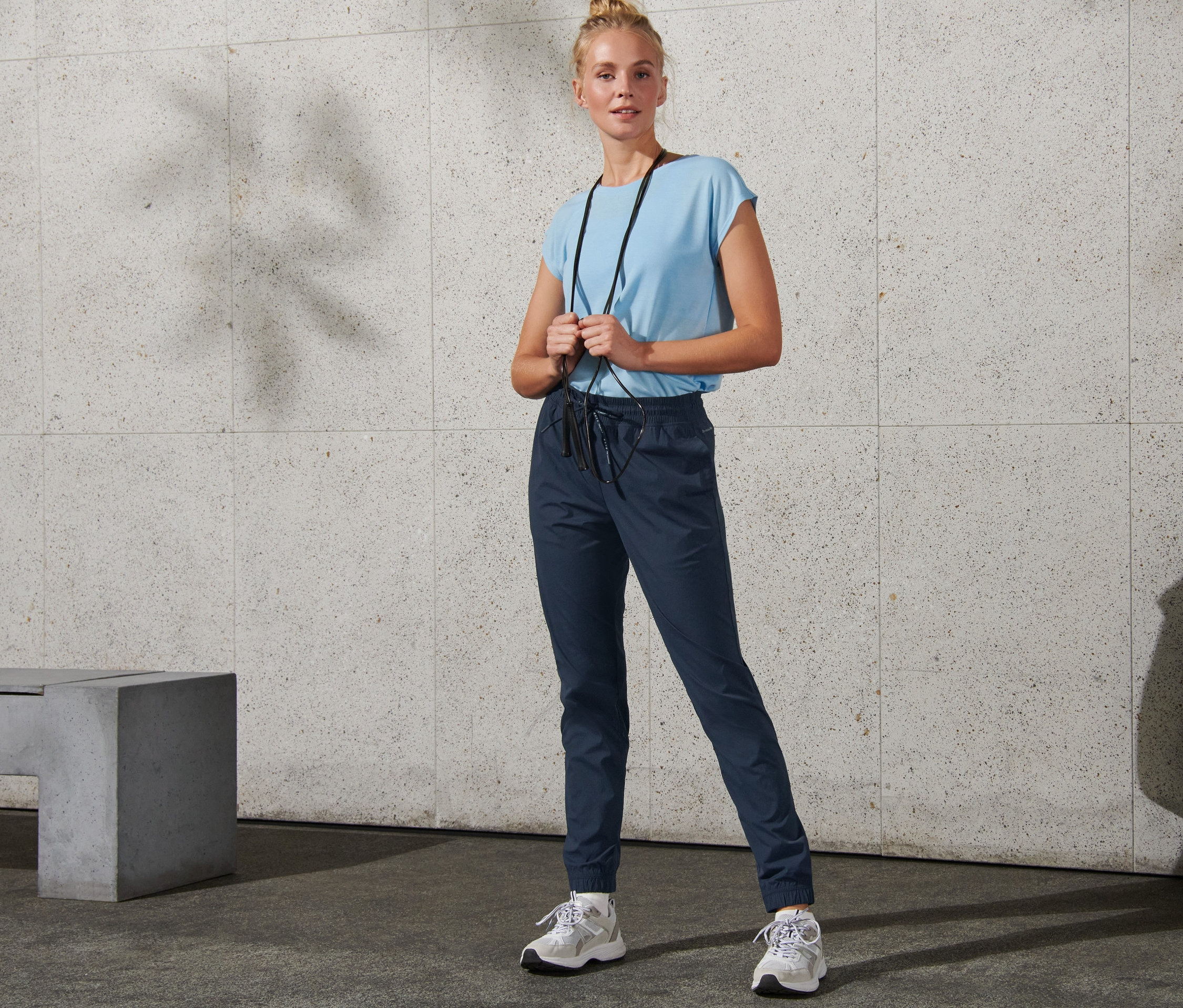 Bir kadın boynunda atlama ipiyle ayakta duruyor ve softshell antrenman pantolonu giyiyor.