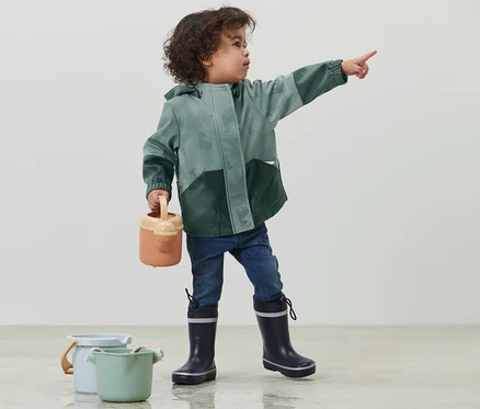 Yeşil Yağmurda Renk Değiştiren Çocuk Softshell Montlu çocuk parmağıyla bir şeyi gösteriyor, elinde sulama kabı, yanında iki kova var.