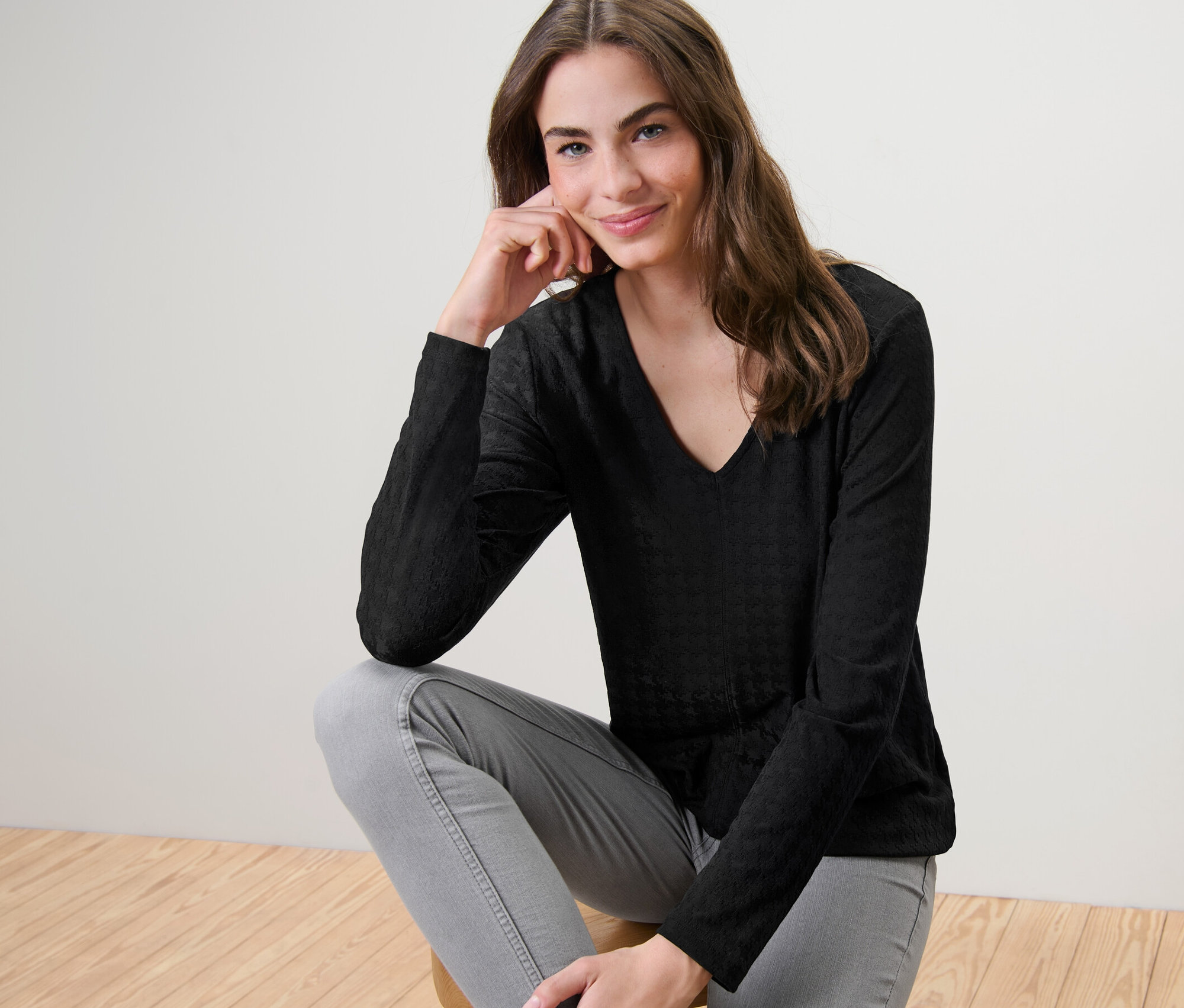 Bir kadın taburede oturuyor ve siyah, dokulu uzun kollu gömlek giyiyor.