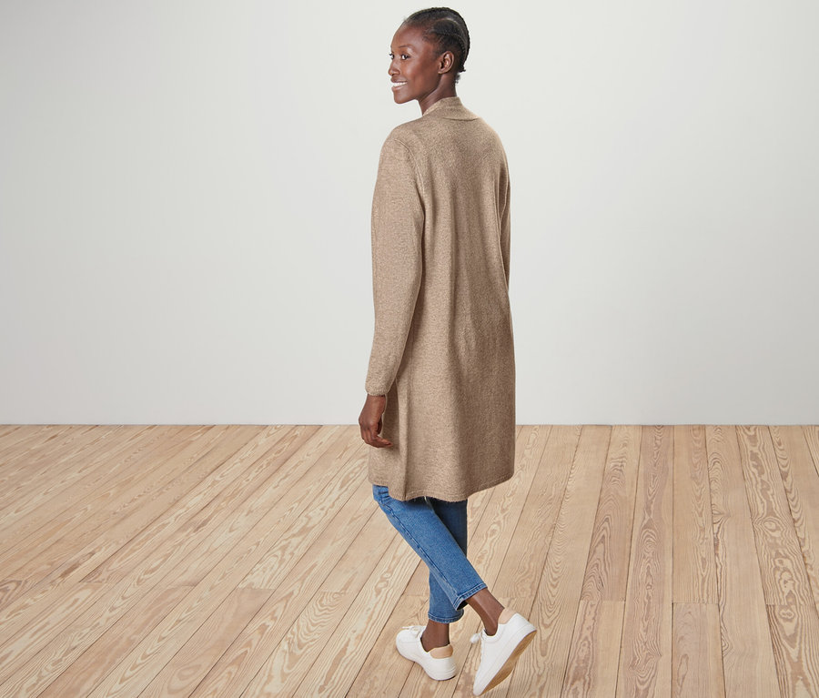 Kadın düğmesiz uzun hırka, mavi kot pantolon ve beyaz ayakkabılarla poz veriyor.