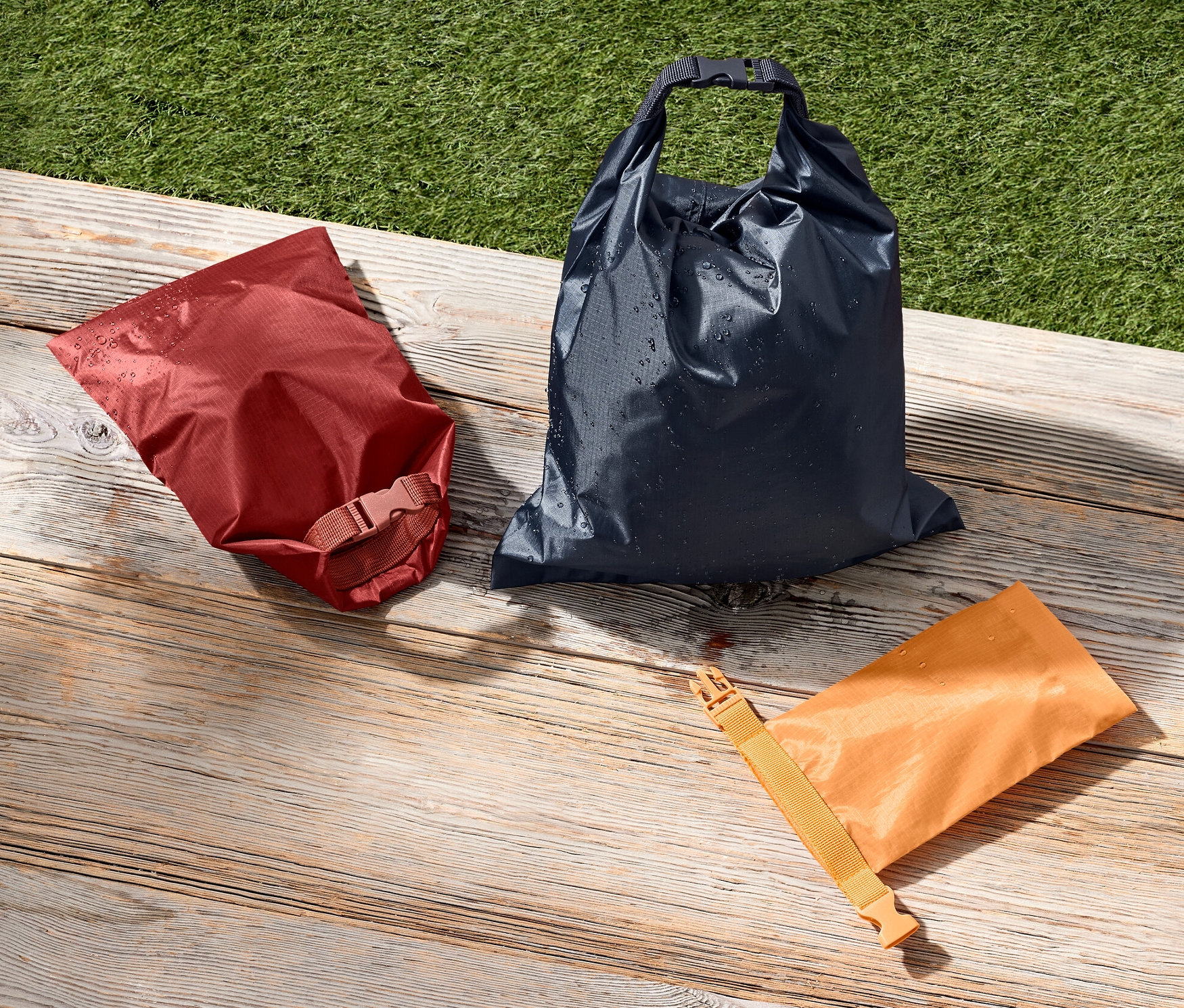 Kırmızı, mavi ve turuncu renklerde üç adet su geçirmez torba, yeşil bir çim alanın yanında ahşap bir yüzey üzerinde duruyor.