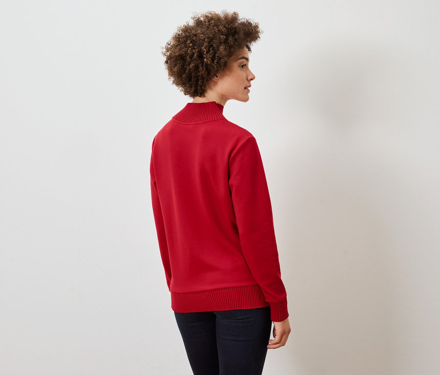 Kıvırcık saçlı bir kadın yan duruyor, kırmızı bir kazak ve koyu mavi kot pantolon giyiyor.