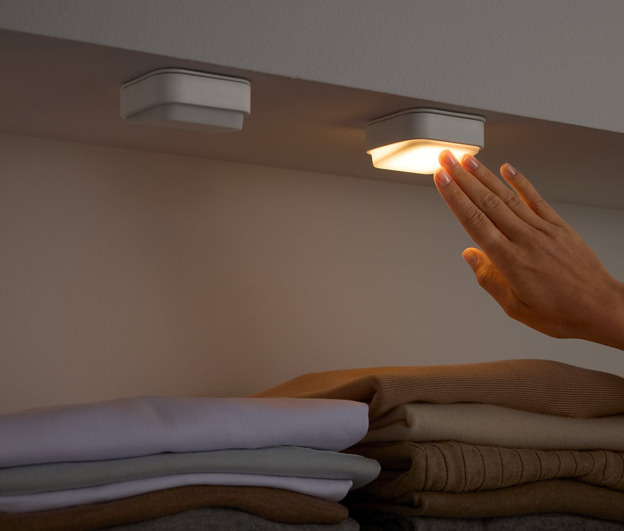 Bir el, katlanmış giysilerin üzerindeki bir rafın altındaki kare bir ışığa dokunuyor.