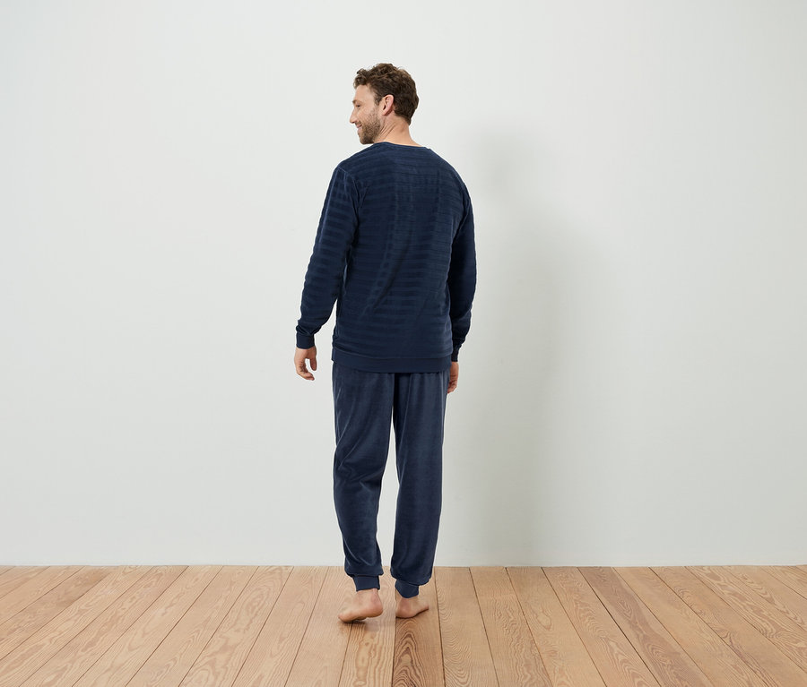 Lacivert çizgili pijamalı bir adam, ahşap zeminde kameraya arkası dönük duruyor.