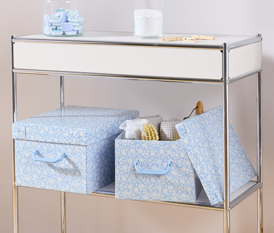 Bir rafta kapaklı iki mavi saklama kutusu ve içinde beyaz havlular ve fırça bulunan kapaklı bir saklama kutusu bulunur.