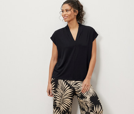 Kadın ayakta duruyor, üzerinde siyah V yakalı bluz ve yaprak desenli pantolon var.