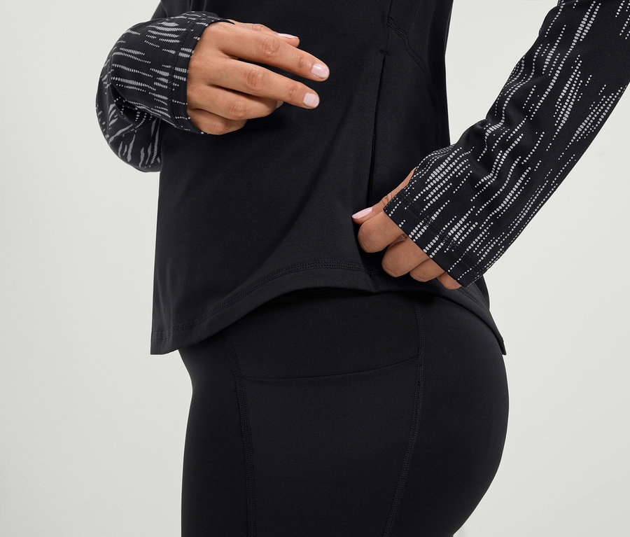 Kadın, siyah uzun kollu bir spor üstü ve siyah taytın yan cebine uzanıyor.