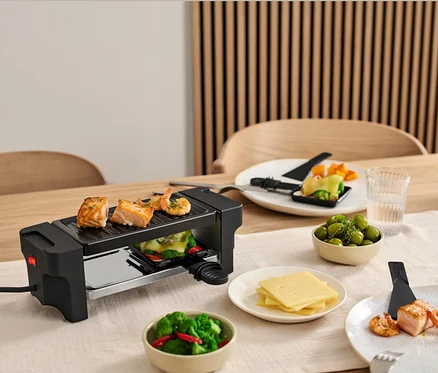 Masa üzerinde 2 Kişilik Raclette Izgara, ızgarada somon ve karidesler var. Malzemelerle dolu tabaklar ızgaranın etrafında duruyor.