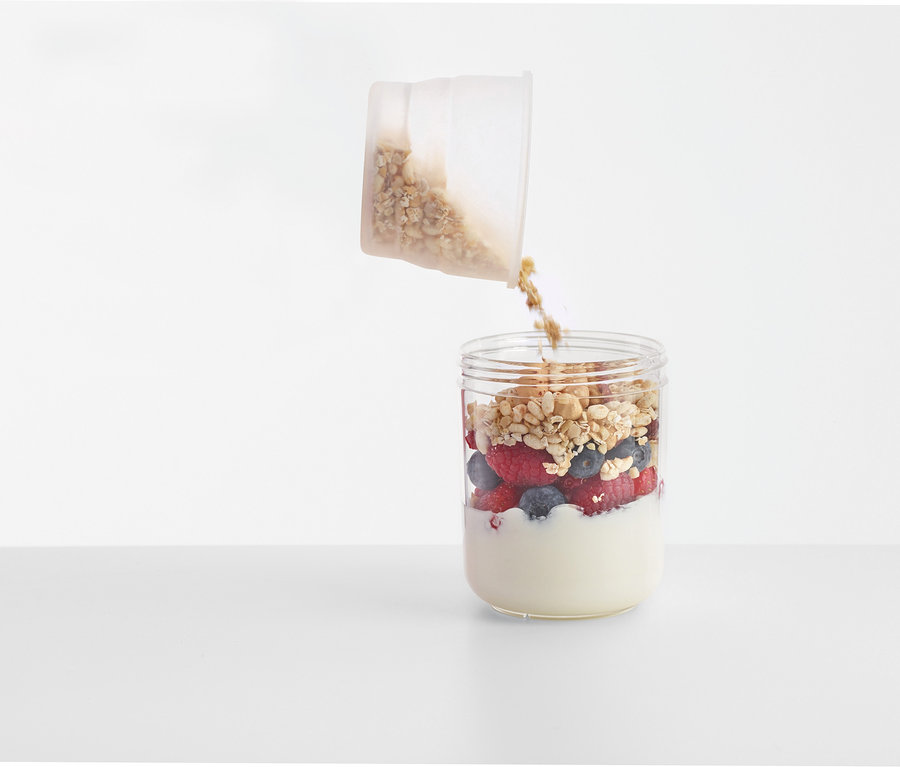 Taşınabilir Atıştırmalık Kutusundan yoğurt ve meyvelerle dolu bir bardağa müsli dökülüyor.