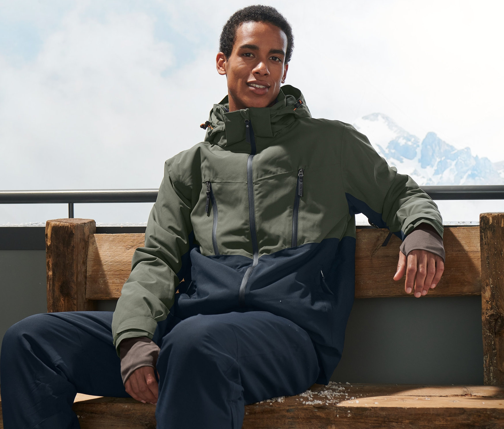 Adam bankta oturuyor, haki kayak montu ve kayak ayakkabıları giyiyor.