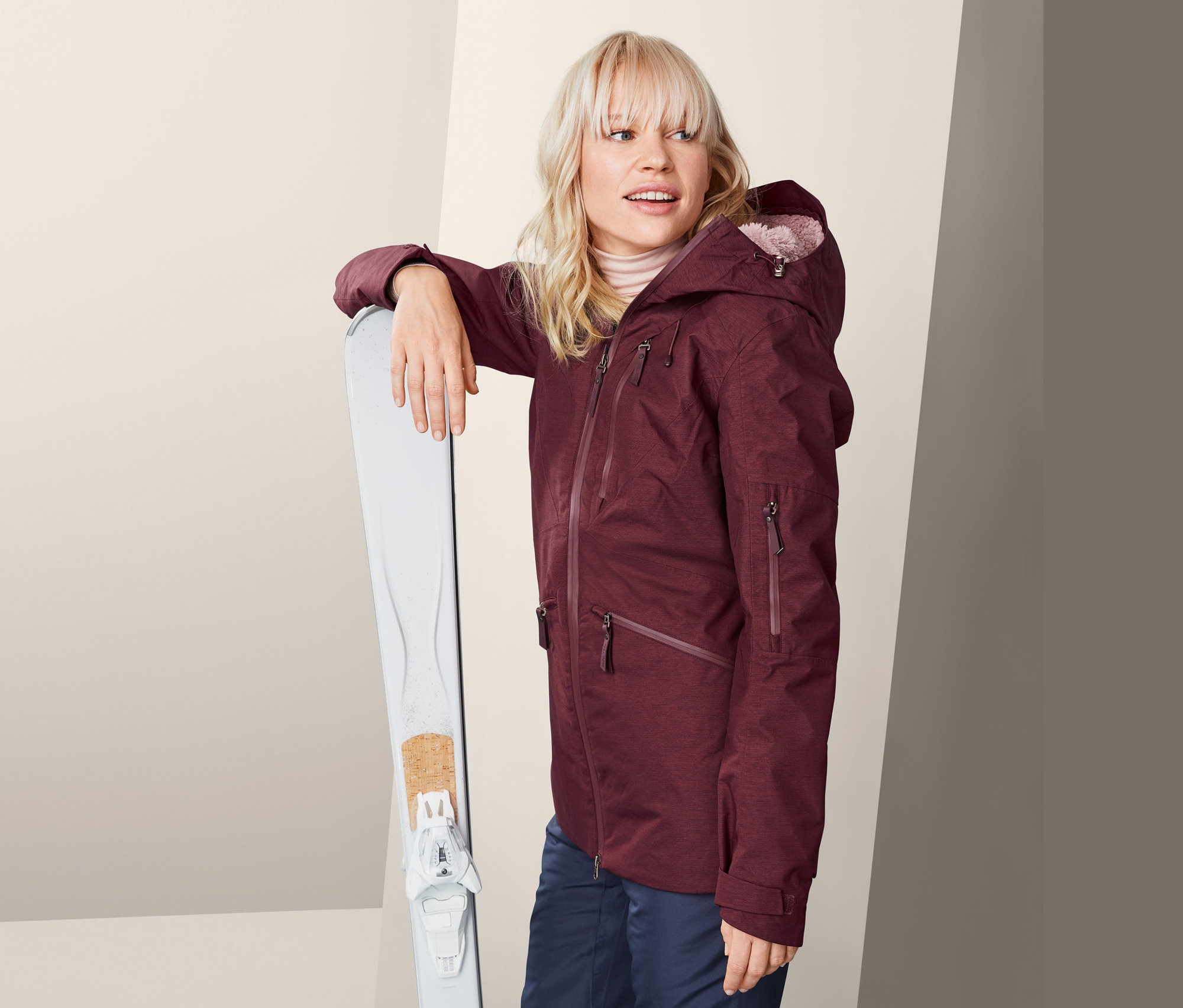 Bordo kayak montlu sarışın bir kadın beyaz kayakların yanında duruyor.